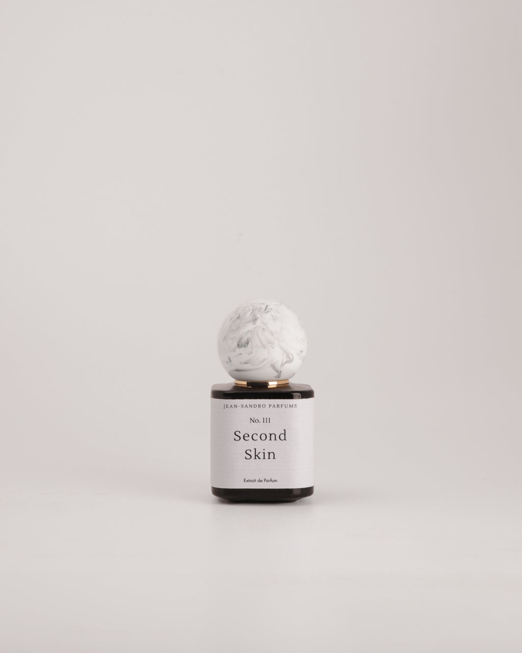 A small cylindrical bottle with a white label reading 'Second Skin' and 'Gentle Handmade Perfumes' near the top, topped with a round white marble-like cap, centered on a light gray background.