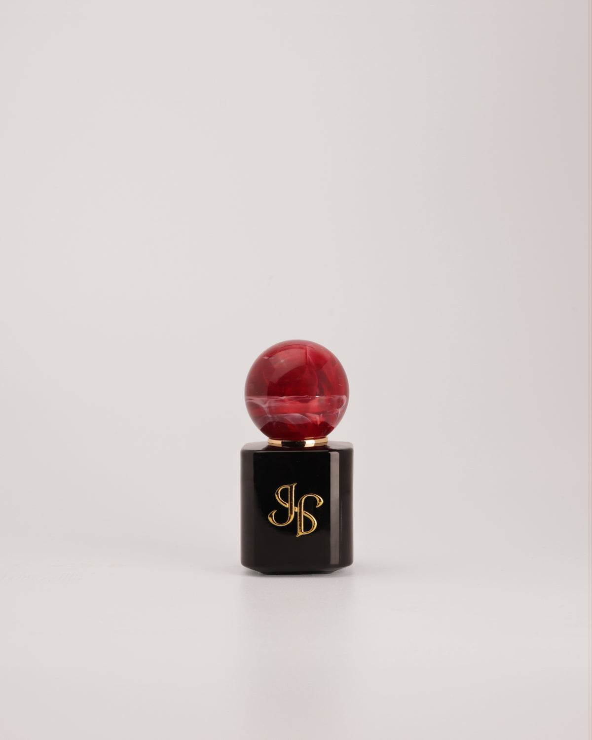 A cosmetic product with a glossy black hexagonal base featuring a gold monogram logo, topped with a round, marbled deep red cap, set against a plain light gray background.