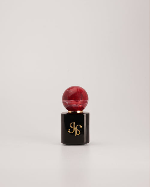 A cosmetic product with a glossy black hexagonal base featuring a gold monogram logo, topped with a round, marbled deep red cap, set against a plain light gray background.