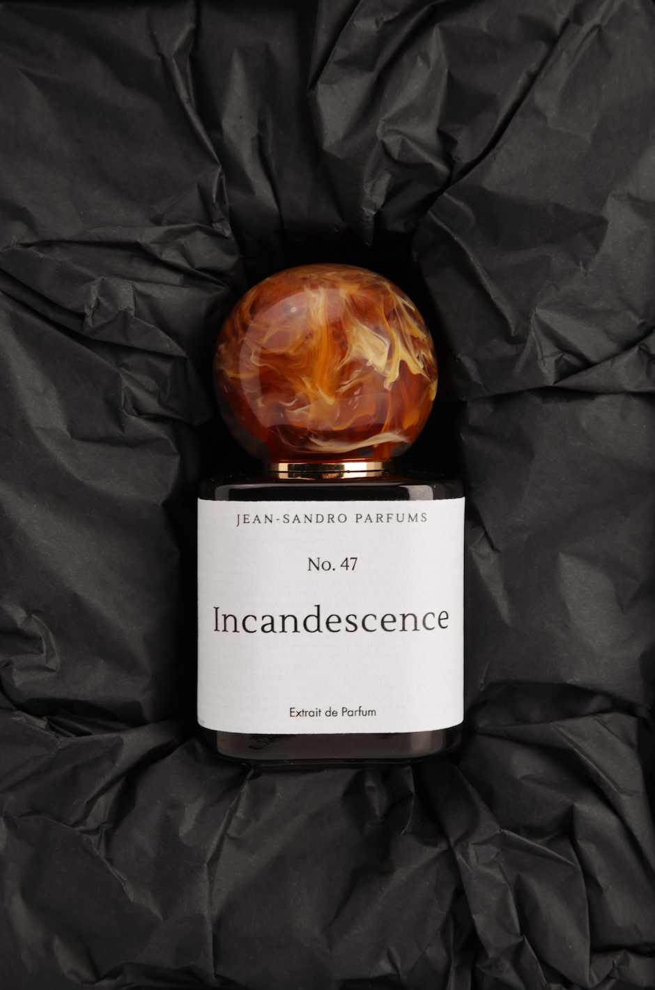 Jean-Sandro Incandescence No. 47 Eau de Parfum bottle with a round amber glass cap, resting on a bed of crumpled black tissue paper.