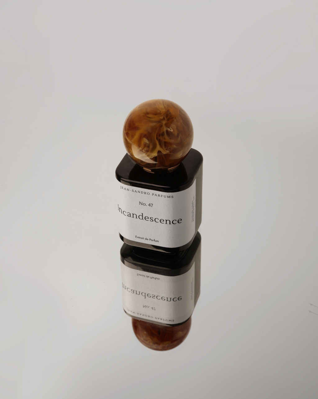 A perfume bottle with a black glass body, a white label reading 'SILVIA SANDBRO KINCIDENCE', and a round amber marbled resin cap, photographed on a light gray reflective surface.