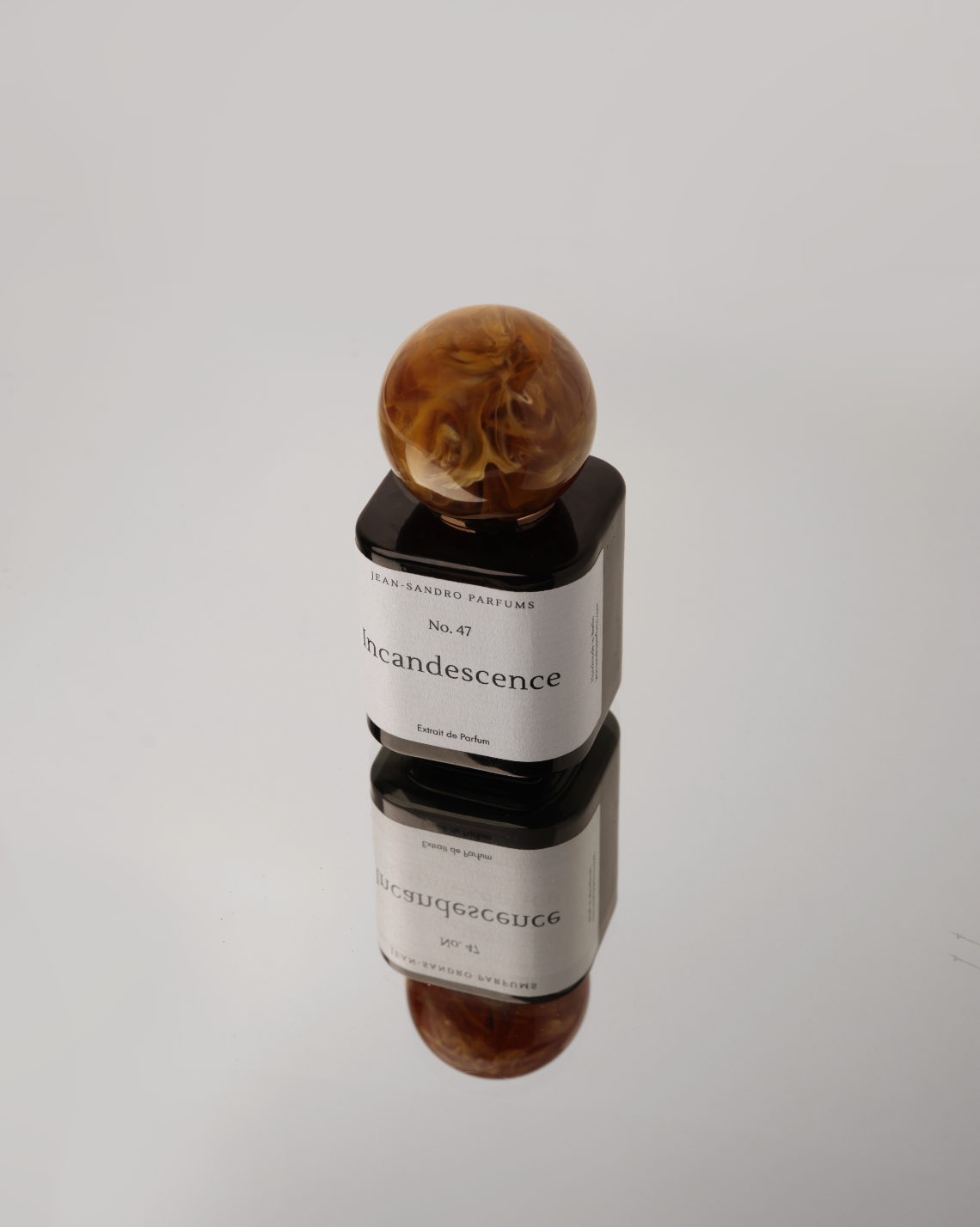 A perfume bottle with a black glass body, a white label reading 'SILVIA SANDBRO KINCIDENCE', and a round amber marbled resin cap, photographed on a light gray reflective surface.