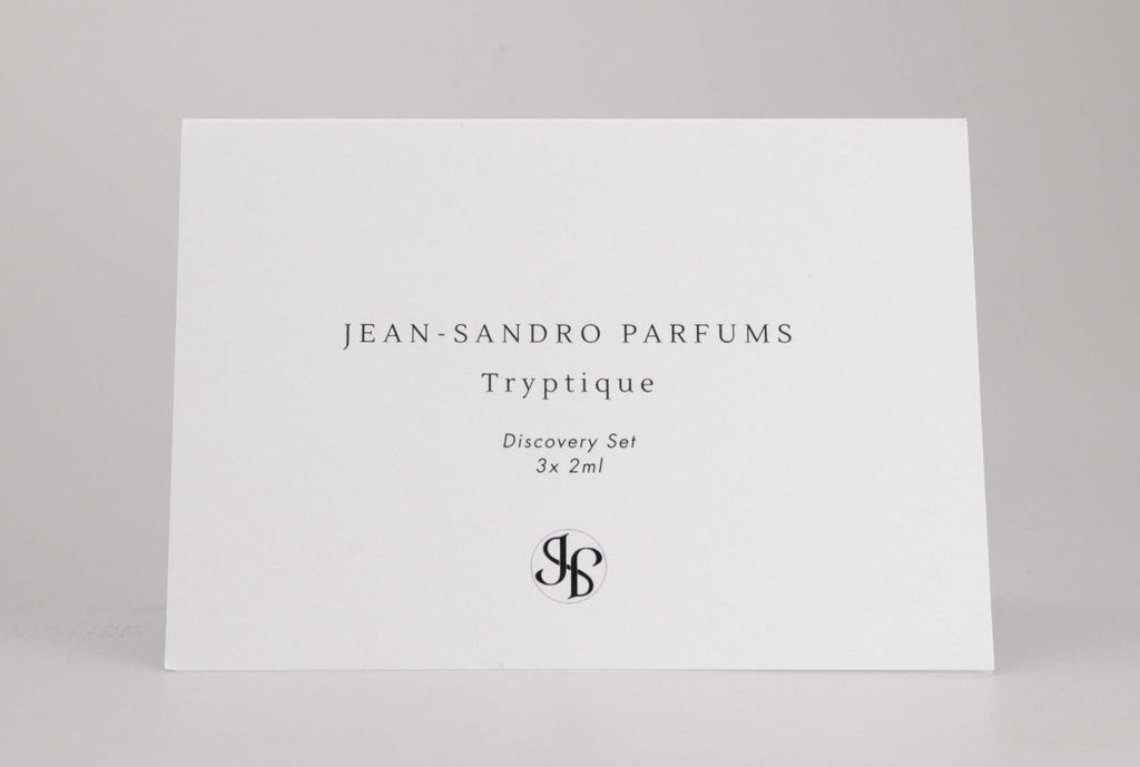 A white rectangular card standing upright on a light gray surface, printed with the text 'JEAN-SANDRO PARFUMS Tryptique Discovery Set 3x2ml' and a monogram logo near the bottom. No person, animal, or real environment is present, so this is Product/Packaging.