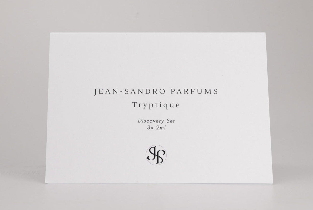 A white rectangular card standing upright on a light gray surface, printed with the text 'JEAN-SANDRO PARFUMS Tryptique Discovery Set 3x2ml' and a monogram logo near the bottom. No person, animal, or real environment is present, so this is Product/Packaging.