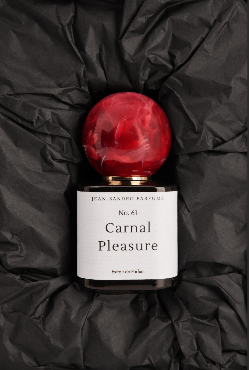 Jean-Sandro Parfums Carnal Pleasure No. 61 perfume bottle with a glossy red spherical cap and a white label, set against a crumpled black background.