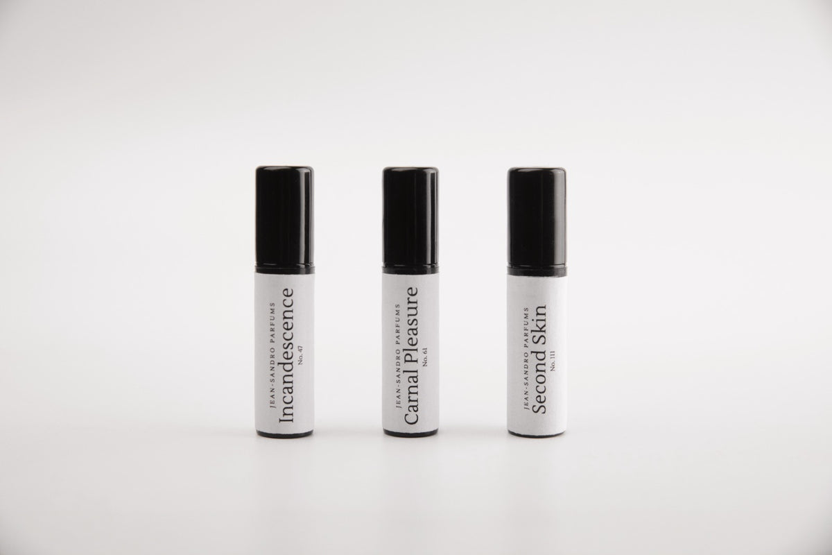 Three small cylindrical cosmetic tubes with black caps and white bodies, arranged side by side on a light gray background. The labels read 'Intocandescence', 'Carnal Pleasure', and 'Second Skin'.