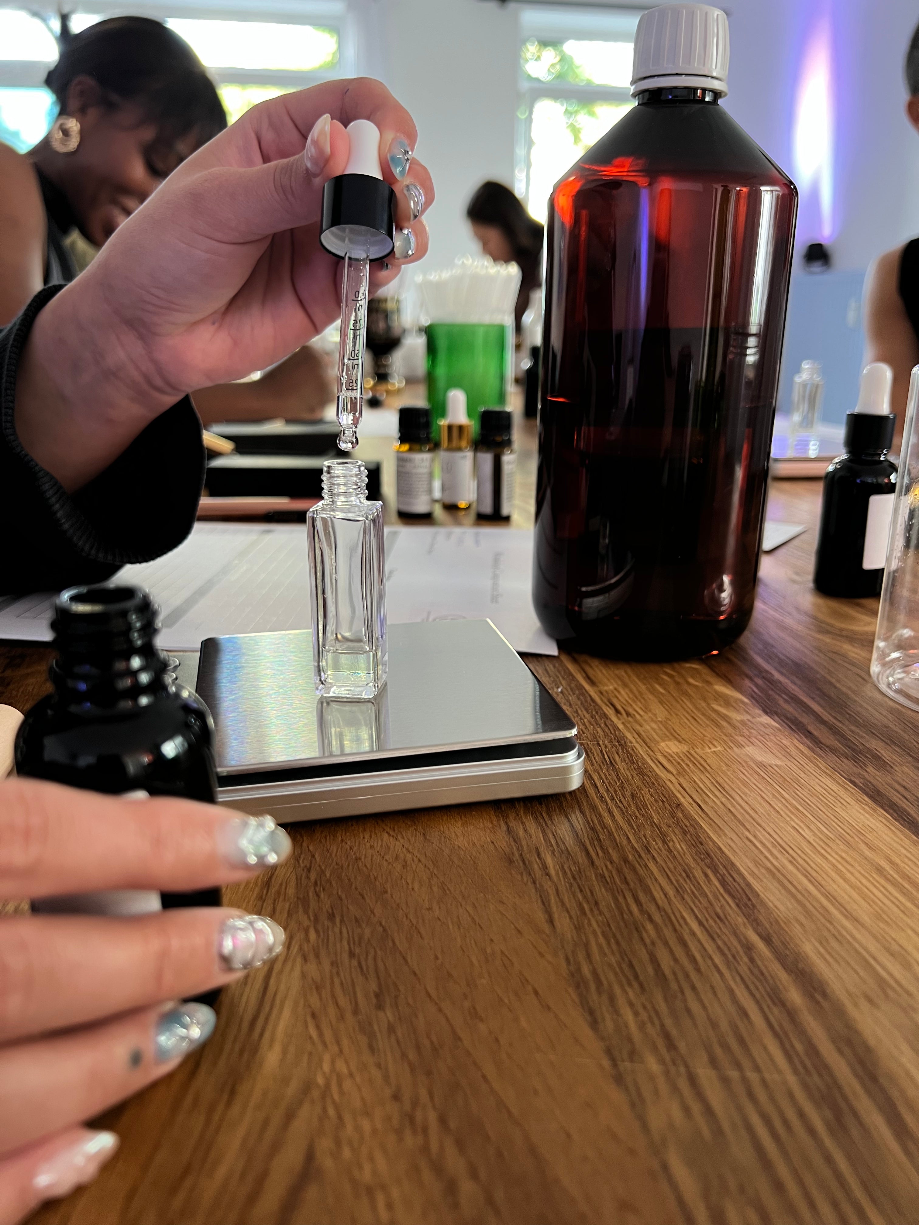 A person's hands holding a glass dropper above a small clear bottle on a digital scale, with larger amber bottles on a wooden table in a bright workshop or studio setting.