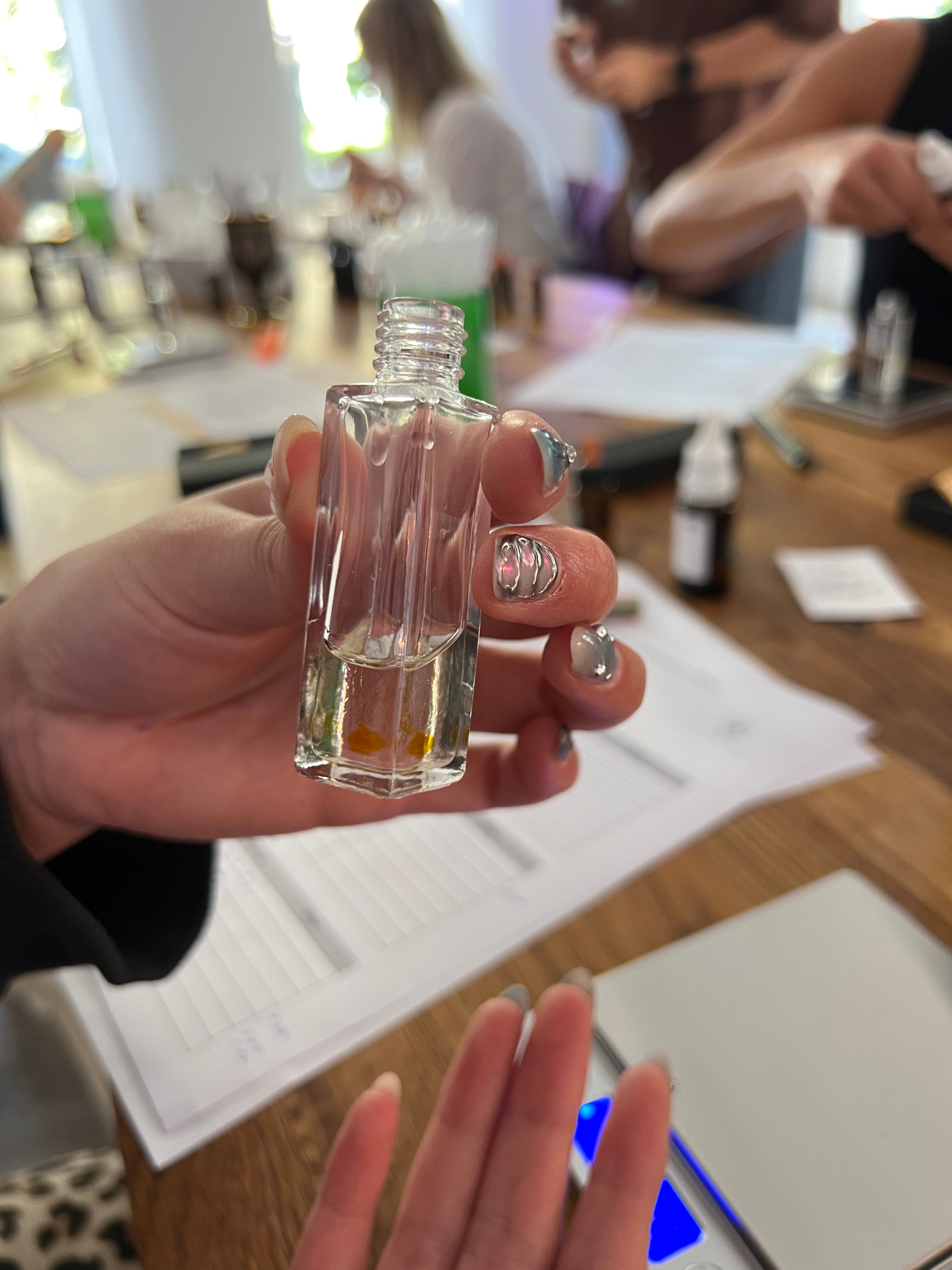 A hand with silver glittery nail polish holds a small, clear glass bottle with a ribbed square shape and an open neck, showing a clear liquid inside. The background is a blurry workshop or studio setting with a wooden table, papers, and other people.